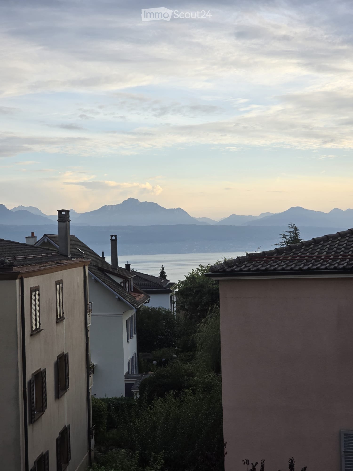 View of residential buildings with tiled roofs and chimneys, overlooking a body of water and distant mountains.