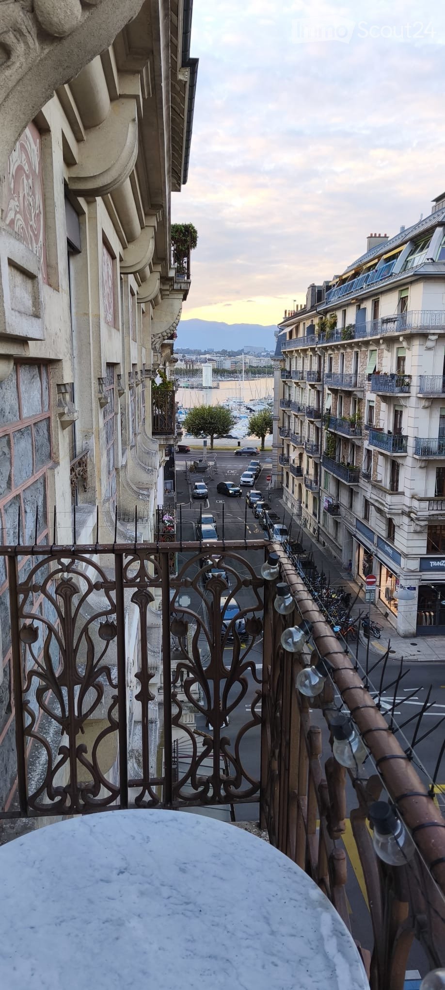 Balcony overlooking a street lined with buildings, cars, and a harbor with boats in the distance.