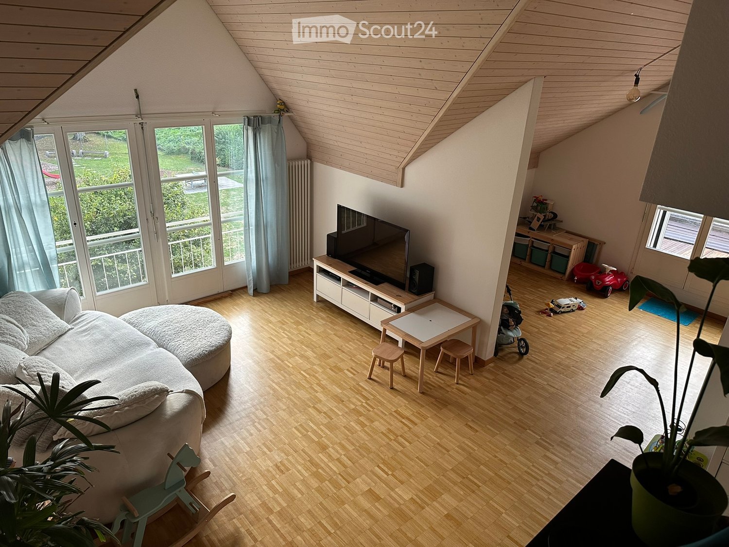 Bright living room with wooden floors, large windows, and a balcony. Features a round sofa, coffee table, small table with stools, TV, and children's toys.