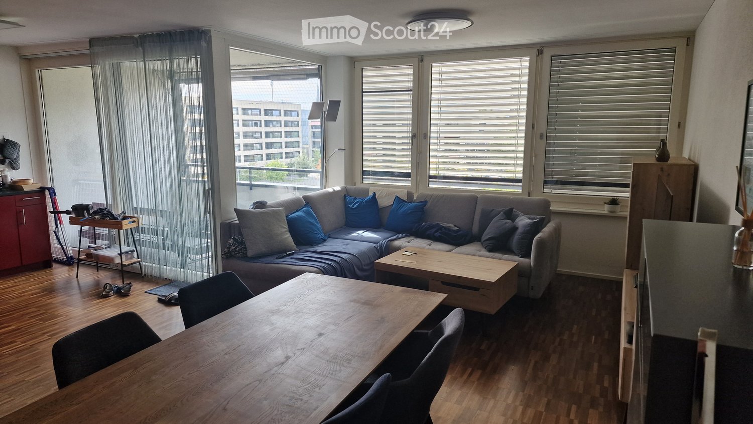 Modern living room with a grey sectional sofa, wooden coffee table, dining table with chairs, and large windows with blinds. The room features a wooden floor and a view of a cityscape.
