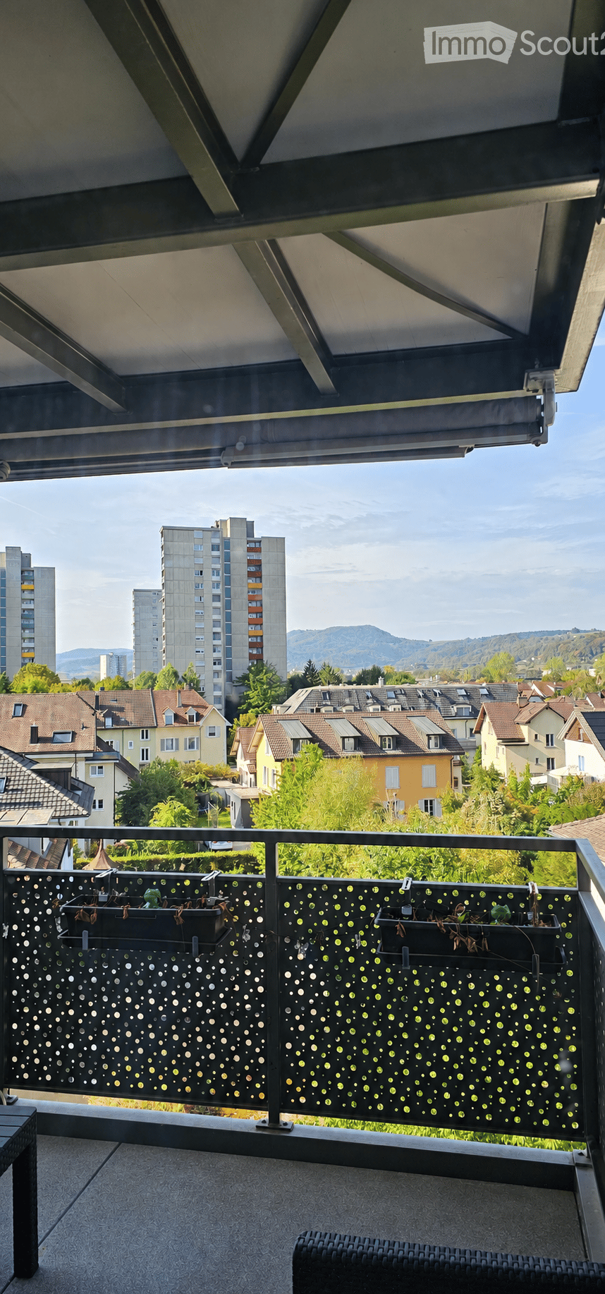 Balcony with modern railing, potted plants, and outdoor furniture overlooking a cityscape with residential buildings and distant mountains.
