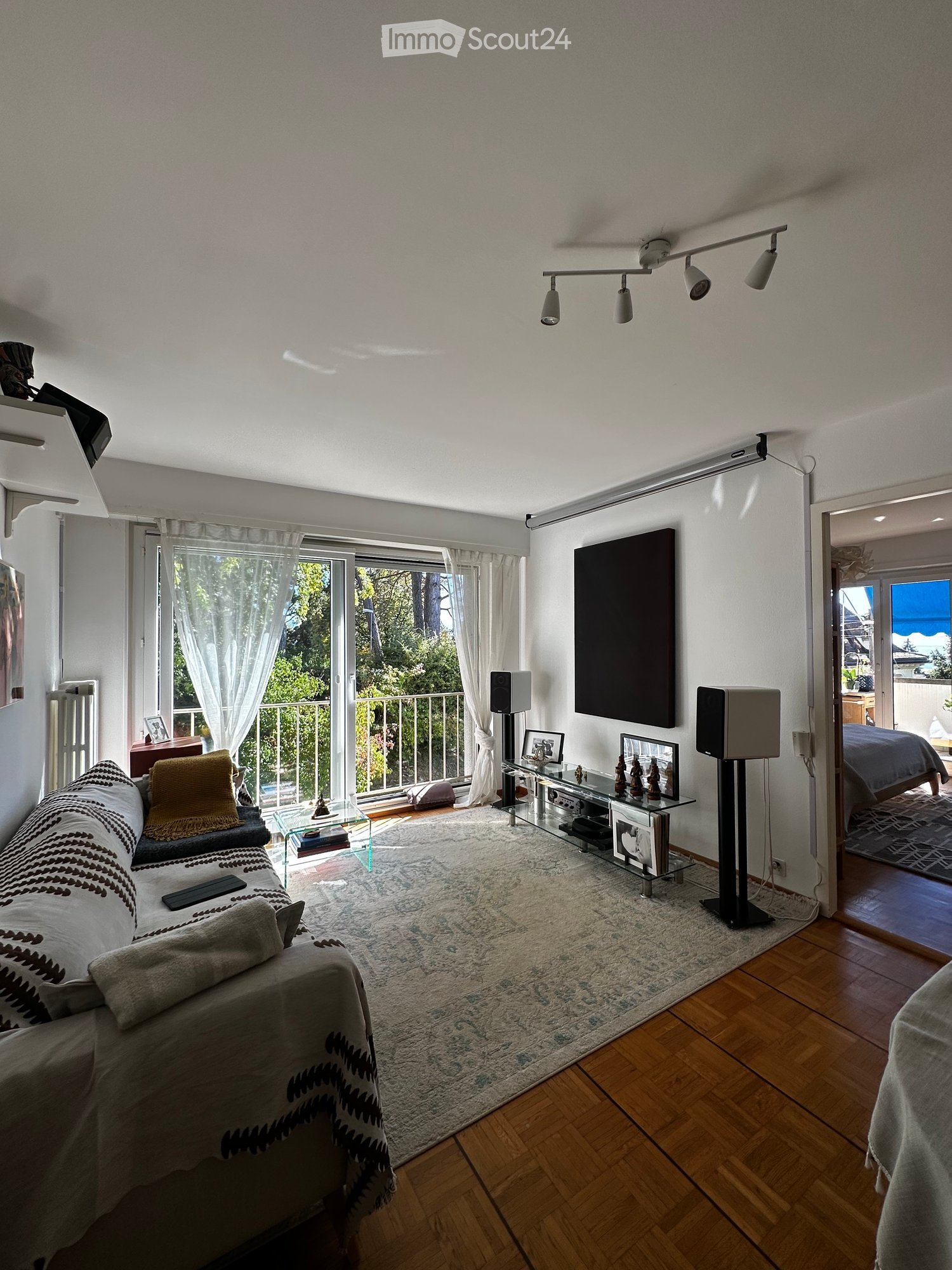 Modern living room with wooden flooring, large windows, a sofa, coffee table, TV stand, speakers, and a view of a balcony.