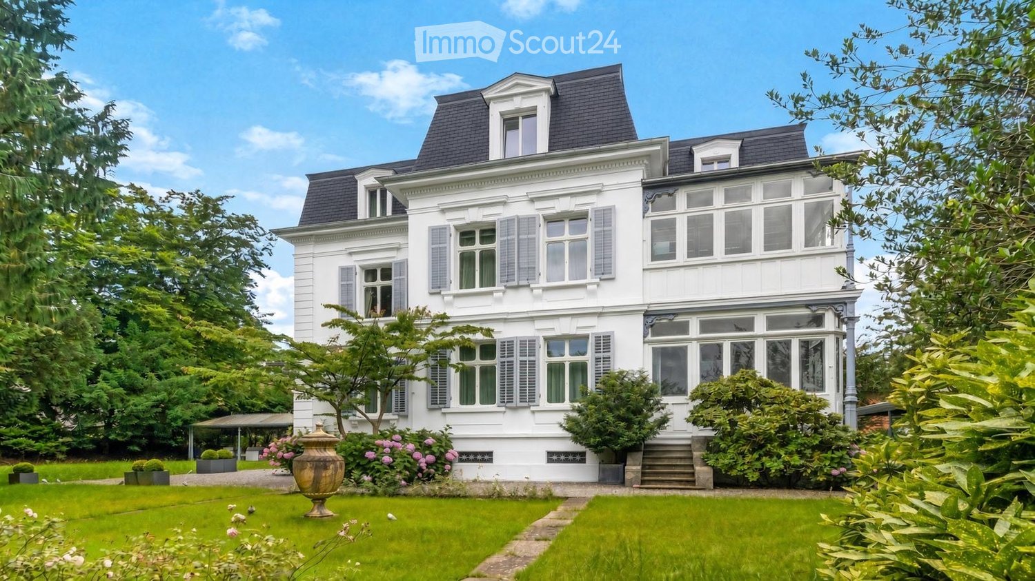 A large, elegant white house with multiple stories, dormer windows, and a well-maintained garden.