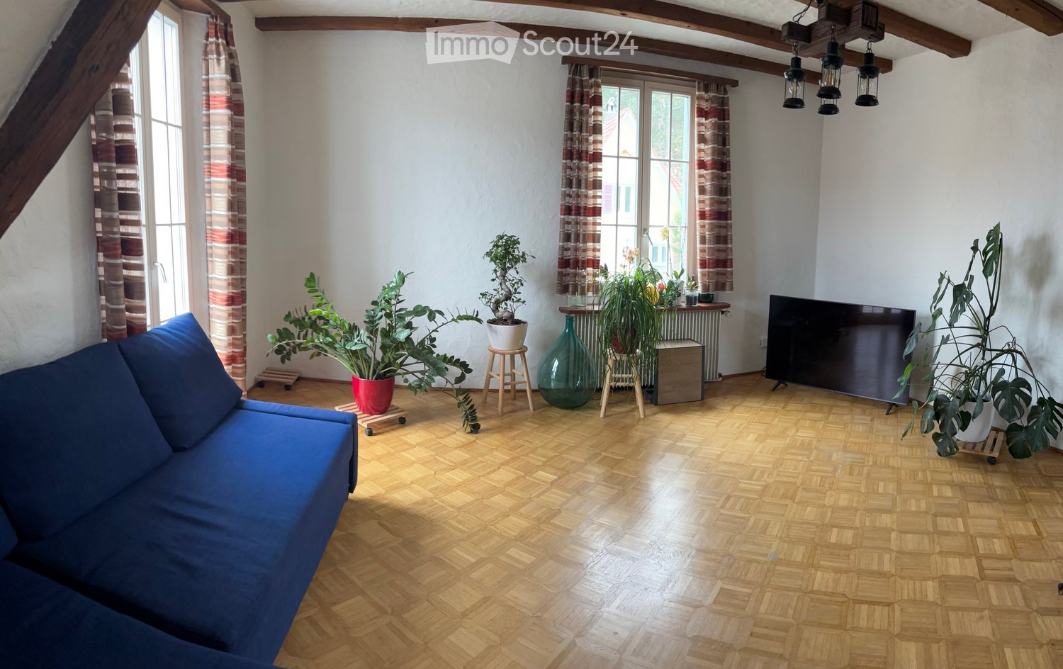 Living room with wooden herringbone flooring, blue sofa, television, potted plants, and decorative vases.