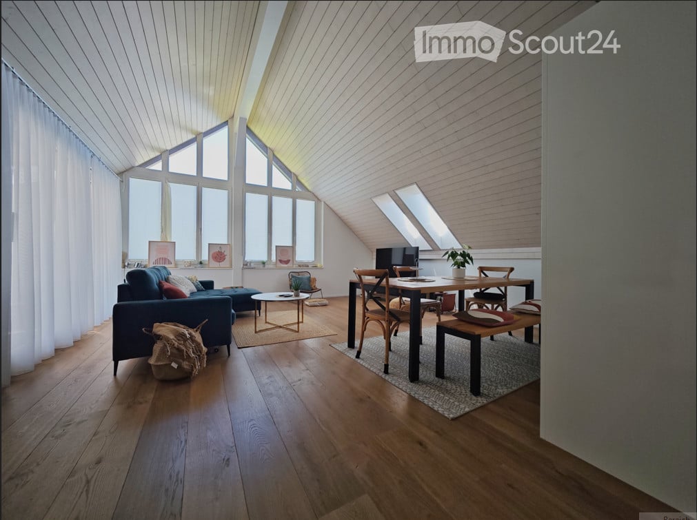 Bright, airy room with wooden floors, large windows, and skylights. Features a blue sofa, coffee table, dining table with chairs, and a small plant on a stool.