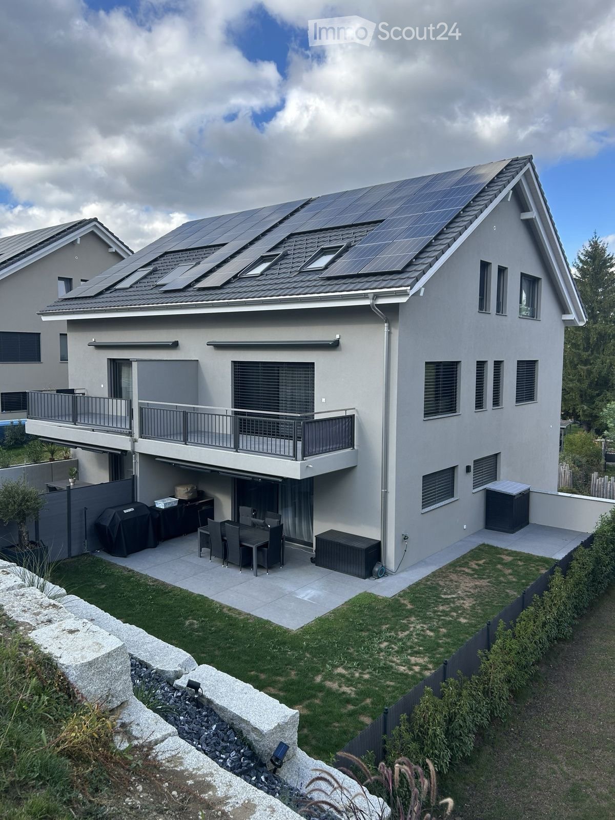 Modern two-story house with solar panels on the roof, large windows, balcony, garage, and outdoor patio with dining set and grill.