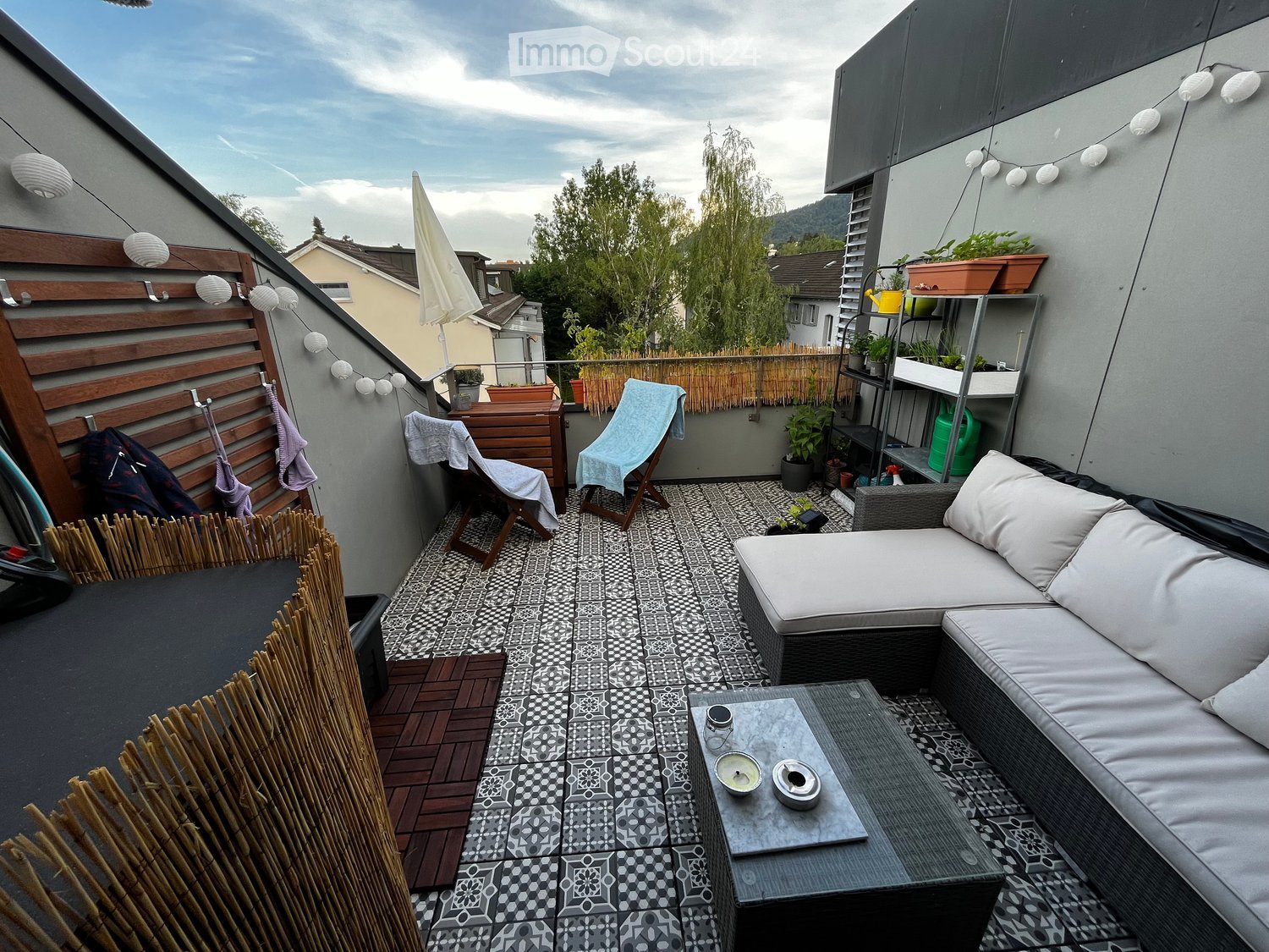 A modern rooftop terrace with a patterned tile floor, a grey sectional sofa, a small coffee table, two wooden chairs with towels, a bamboo screen, and potted plants on shelves.