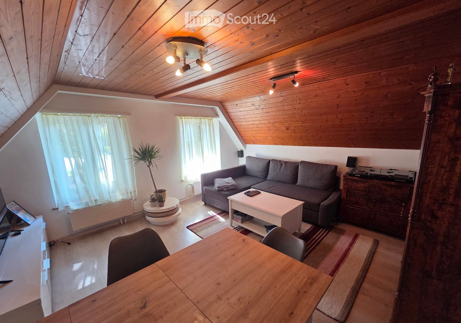 A cozy living room with wooden beams on the ceiling, a grey sofa, a white coffee table, a wooden dining table, and a potted plant near the window.