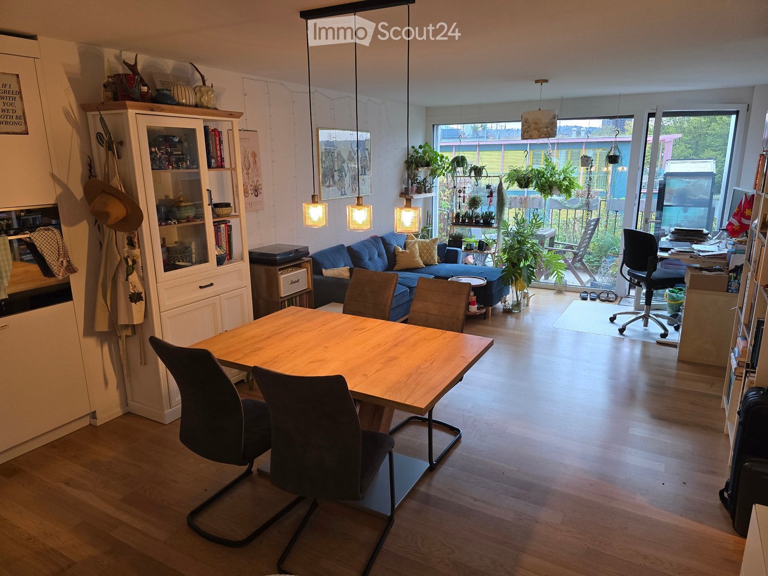 A modern, well-lit living and dining area with a blue sofa, wooden dining table, chairs, bookshelf, and hanging plants. The space features large windows with a view of the garden.