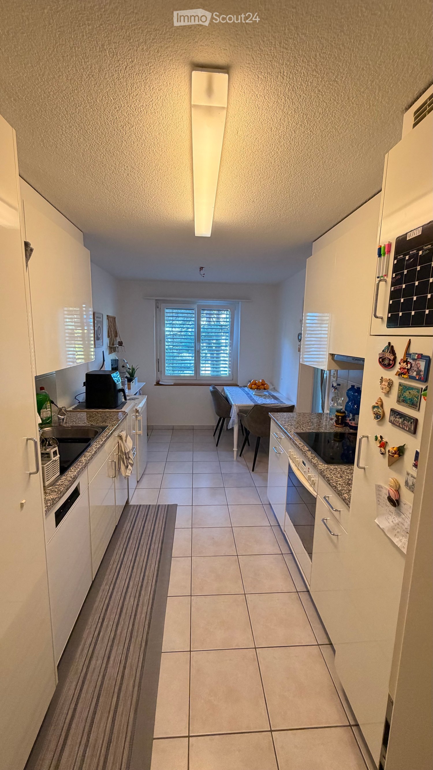 Modern kitchen with white cabinets, granite countertops, and a dining area with a table and chairs. Features include a refrigerator with magnets, a coffee maker, and a calendar on the wall.