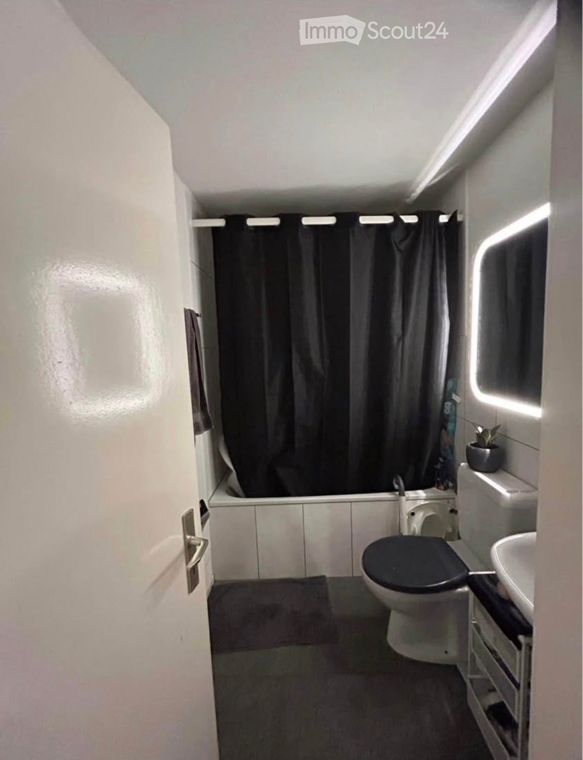 Compact bathroom with a toilet, sink, bathtub, and a small plant on the counter. The space features white tiled walls and a dark grey shower curtain.