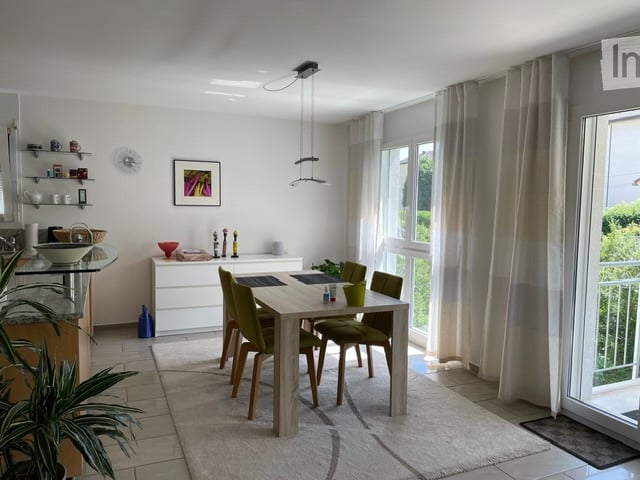 Modern dining area with a light wood dining table and four green chairs, a kitchen island with shelves, and large windows with curtains. A framed picture hangs on the wall, and a potted plant adds a touch of greenery.