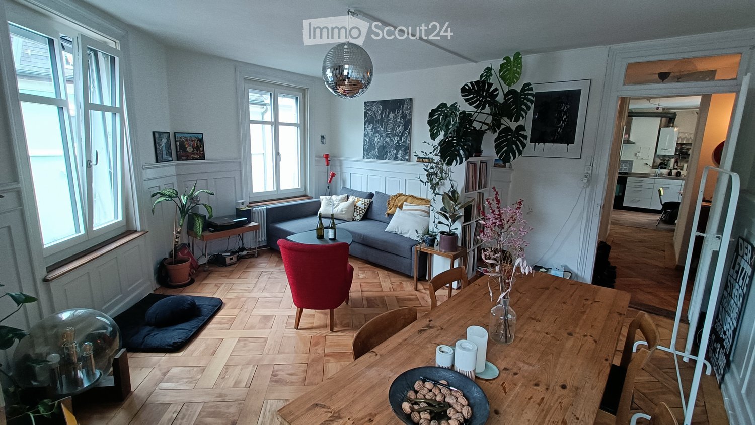 Bright living room with wooden flooring, large windows, a grey sofa with pillows, a coffee table, a red chair, a dining table with chairs, and decorative plants.