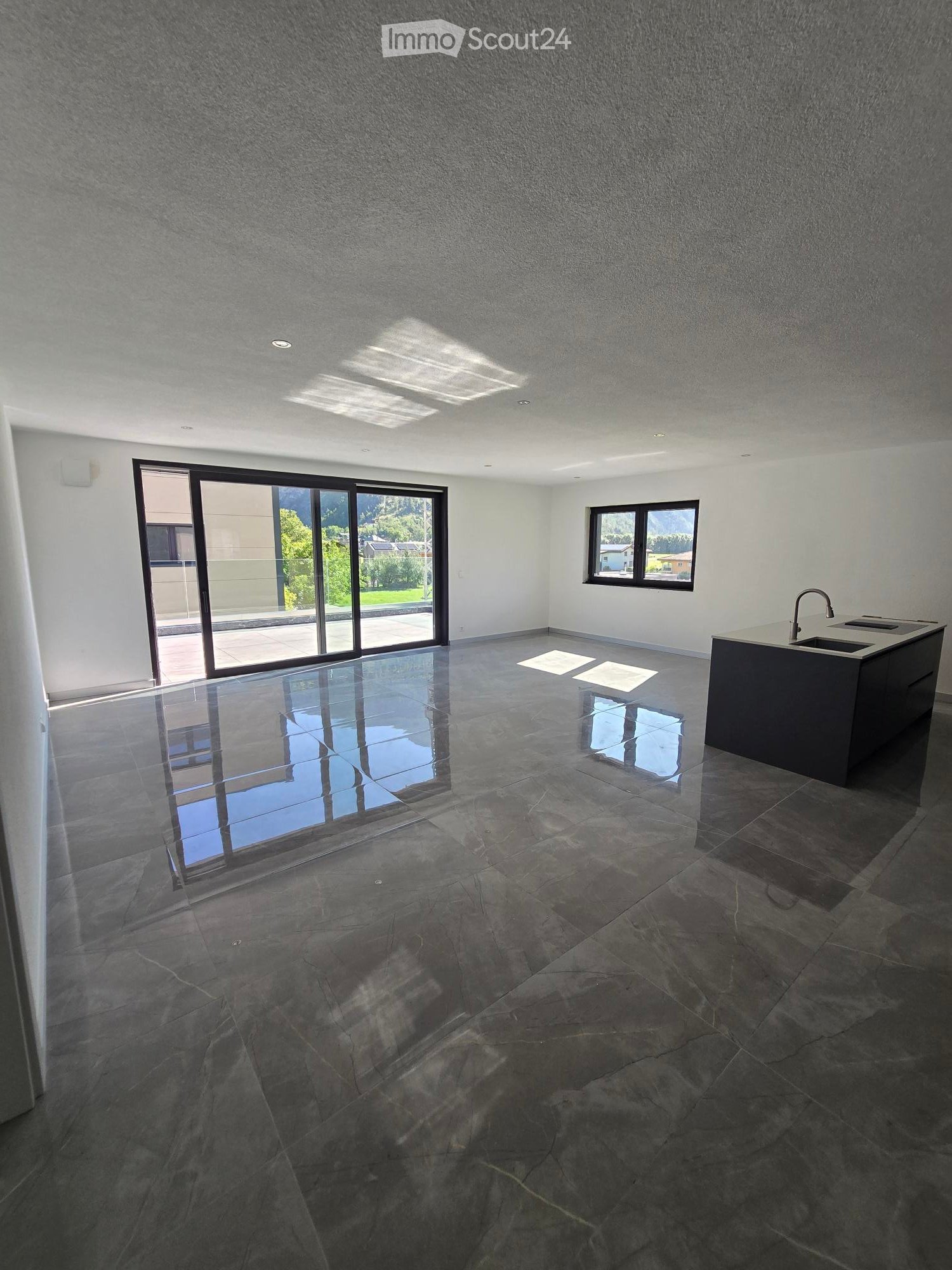 Empty room with dark stone tiles, large windows, and a modern kitchen island with a sink.