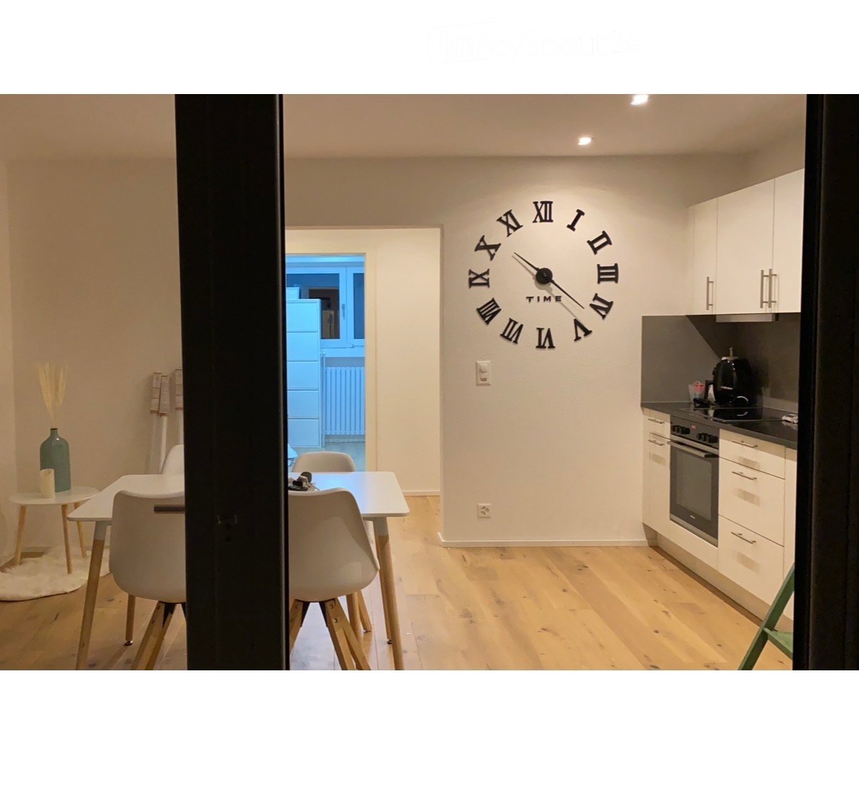 A modern, open-plan living and dining area with a kitchen. The space features a large wall clock, white dining table with chairs, a small side table with a vase, and a kitchen area with cabinets, an oven, and a coffee maker.