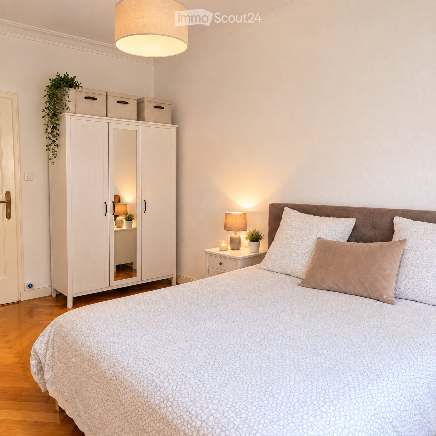 Modern bedroom with a large bed, nightstand, lamp, wardrobe with mirror, and potted plants.