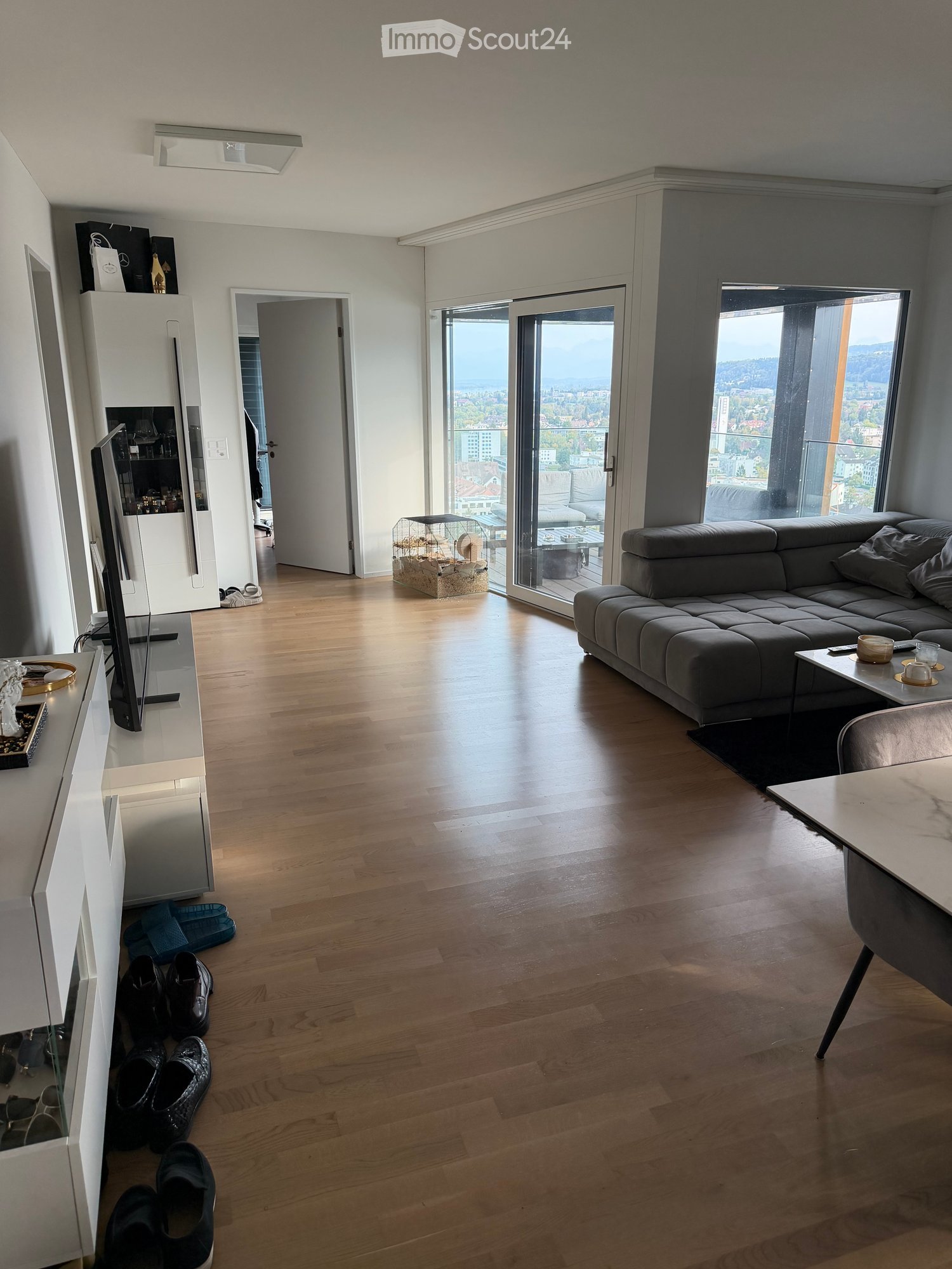 Modern living room with wooden flooring, large windows offering a city view, a grey sectional sofa, a coffee table, a TV stand, and a small table with chairs.