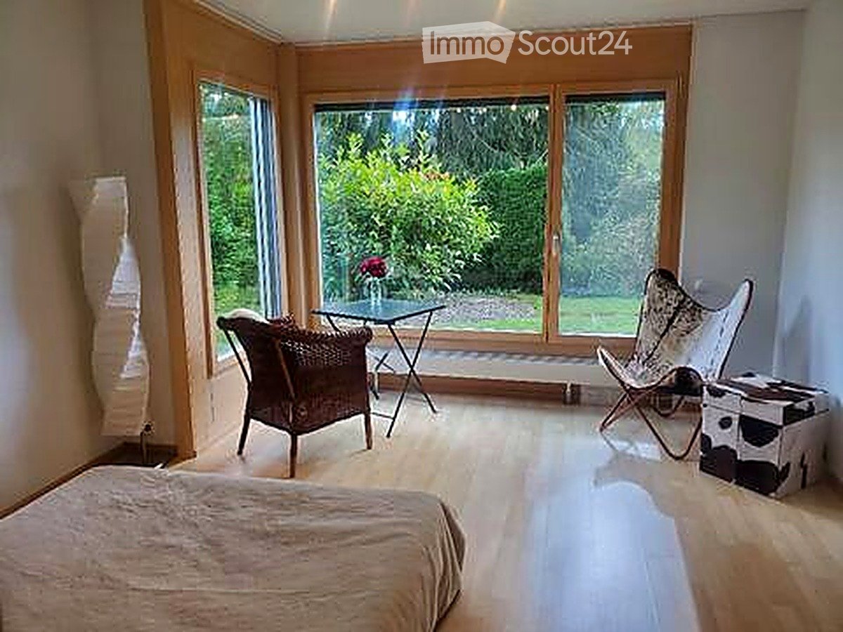 Bright, modern bedroom with large windows overlooking a garden, featuring a bed, wicker chair, small table, and decorative stool.