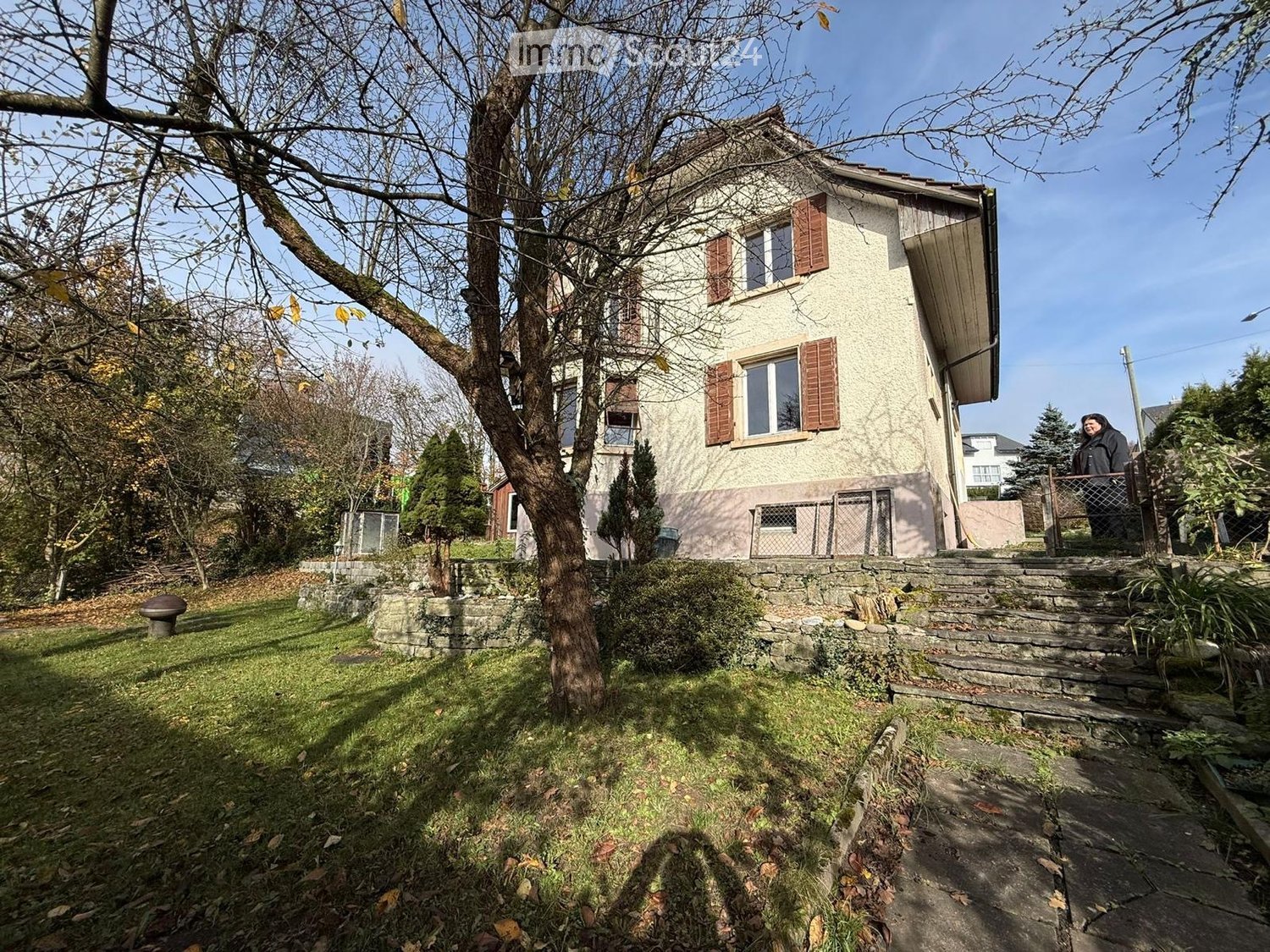 Two-story house with beige siding, red shutters, and a stone pathway leading to the entrance. The house is surrounded by a well-maintained garden with trees, shrubs, and a stone bench.
