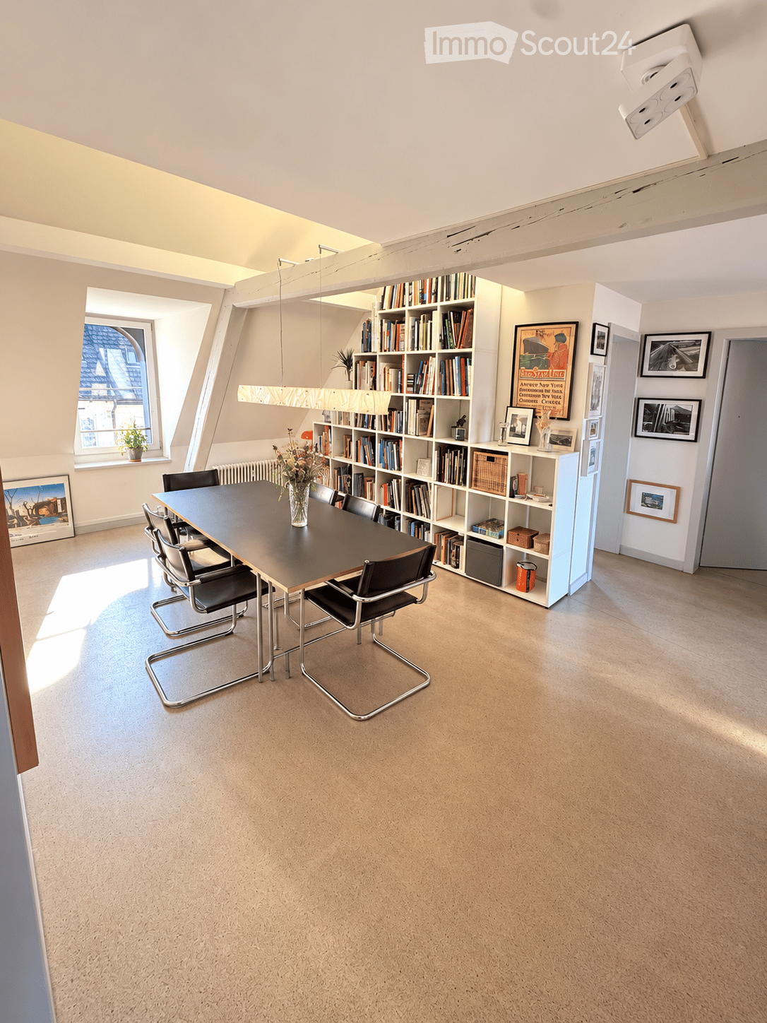 Bright, modern room with a large wooden table surrounded by black chairs, a bookshelf filled with books, and several framed pictures on the walls. Natural light streams in from a large window on the left side.