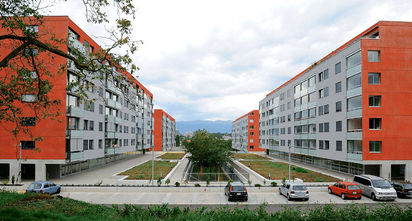 Modern apartment complex with red and grey facades, multiple floors, balconies, and a parking area with several cars.