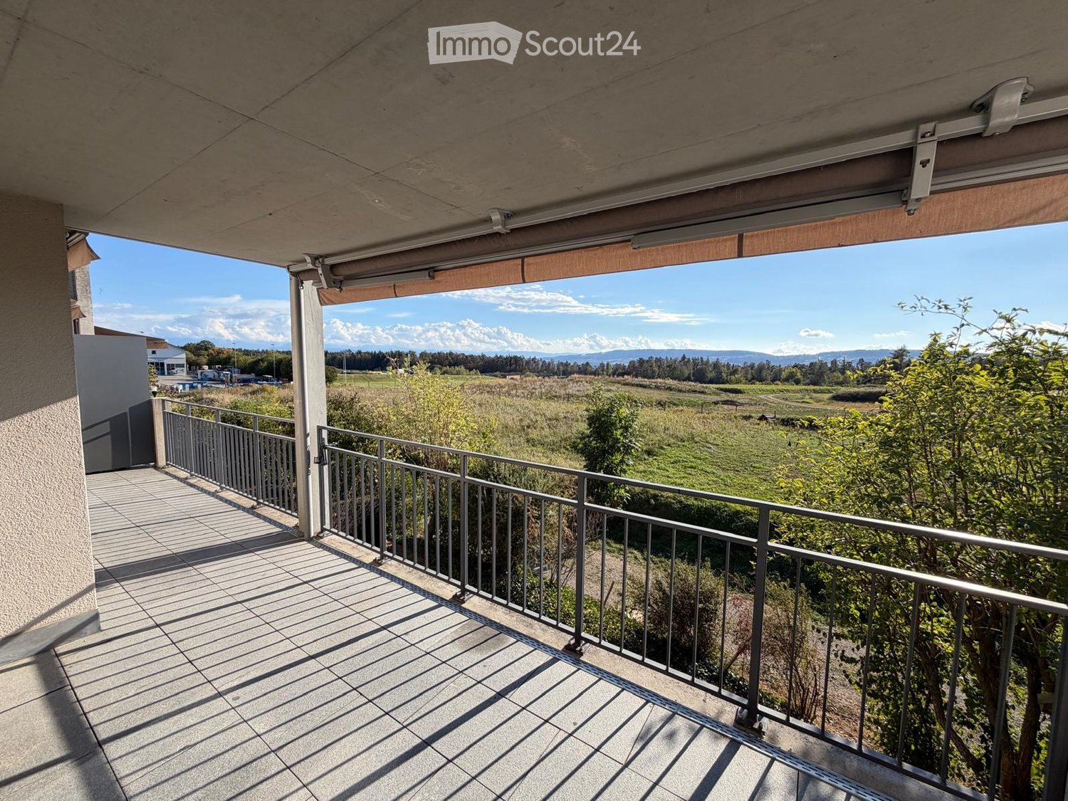 Balcony with tiled floor, metal railing, and a retractable awning, overlooking a green field and distant mountains.
