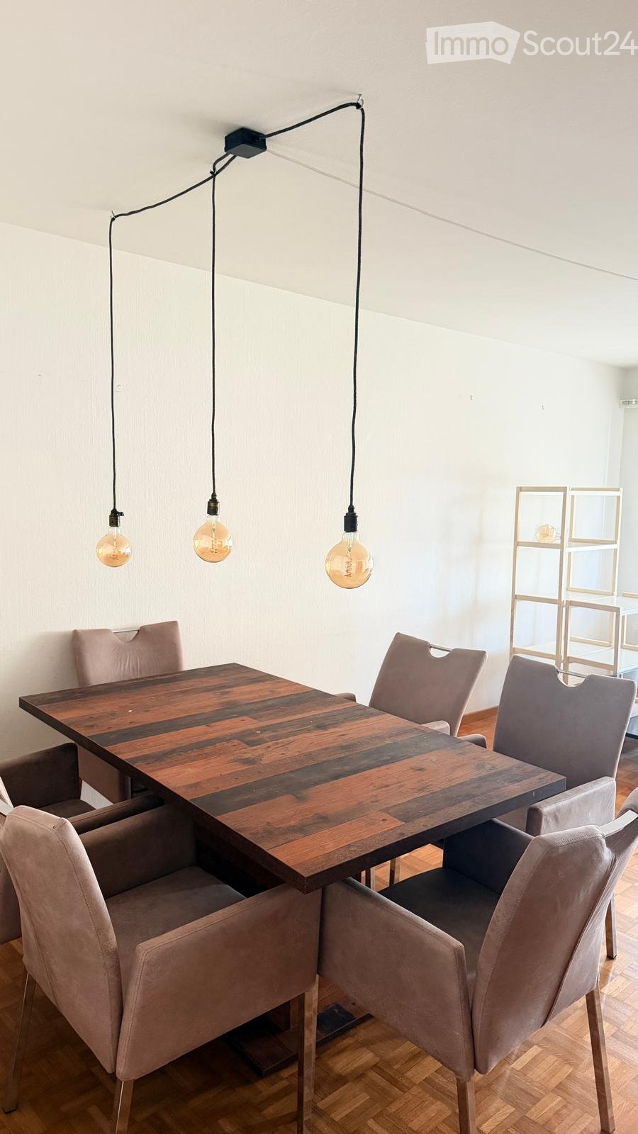 Dining area with a wooden table and six upholstered chairs, pendant lights hanging from the ceiling, and a modern shelving unit in the background.