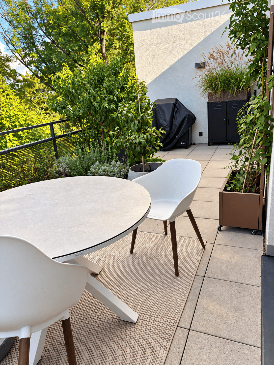 Modern balcony with a round table, two white chairs, potted plants, and a covered grill.