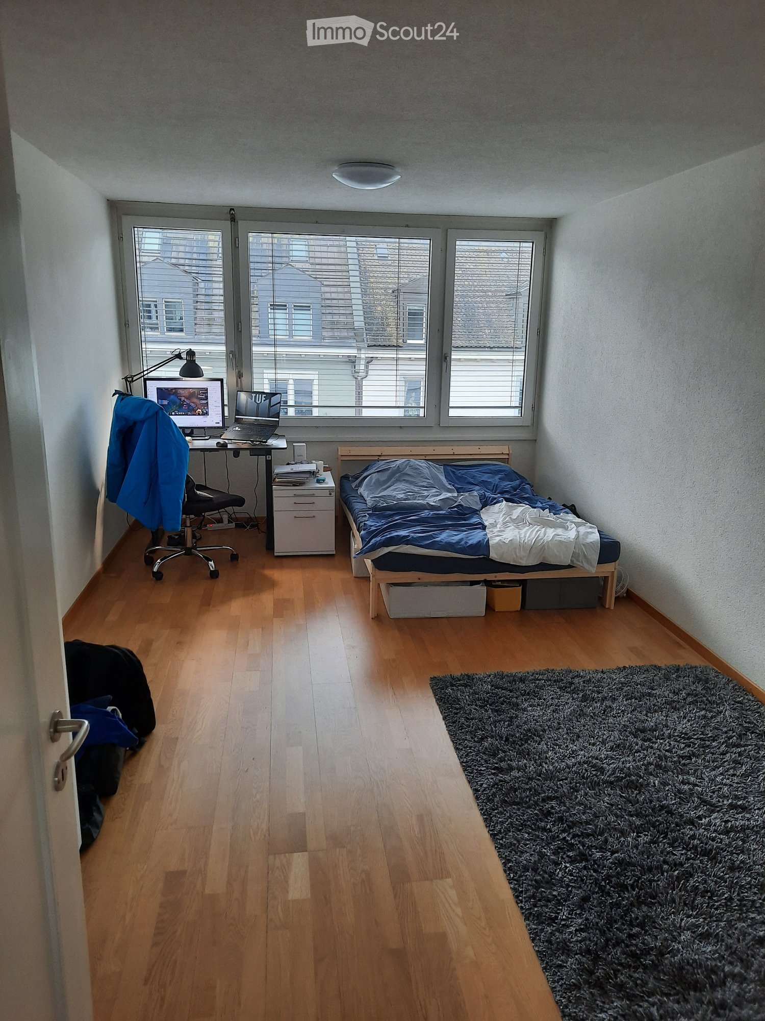 A small, well-lit bedroom with a wooden floor, a single bed with blue bedding, a desk with a laptop and chair, a storage unit, and a large shaggy rug near the bed.