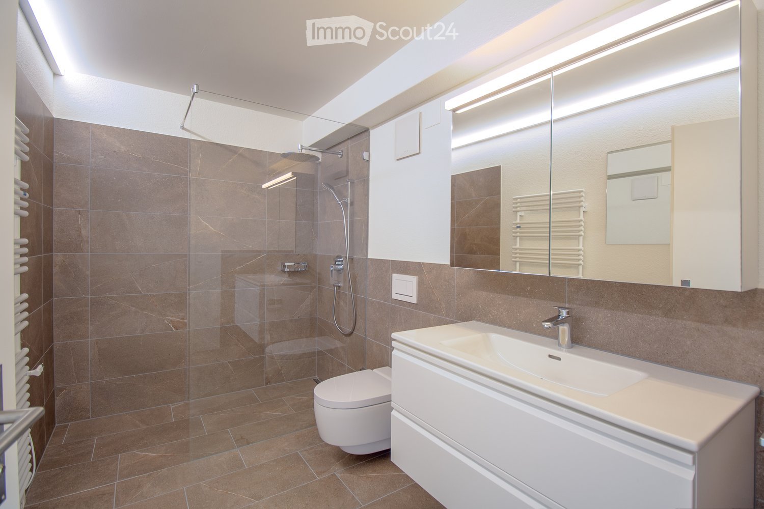 Modern bathroom with brown tiled walls and floor, white sink, toilet, large mirror, and glass-enclosed shower area.