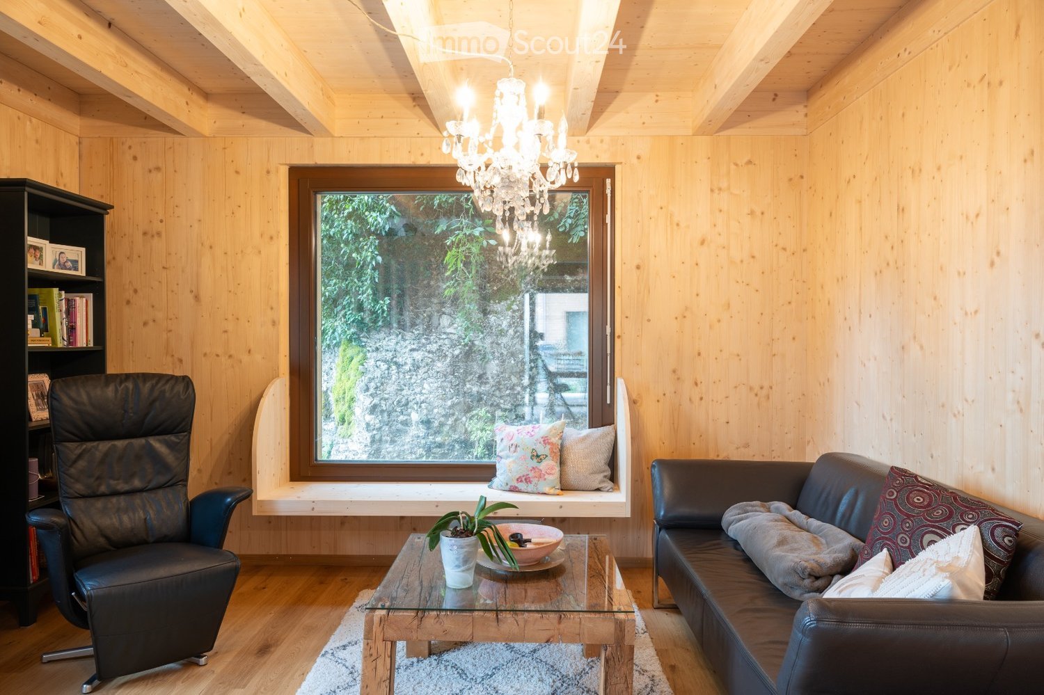 Cozy living room with wooden walls and ceiling, featuring a large window with a view, a leather sofa, a leather armchair, a glass coffee table, a bookshelf, and a chandelier.