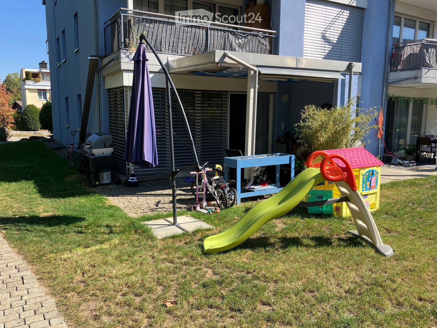 Garden area with a slide, toy house, bicycle, table, chair, umbrella, and grill, adjacent to a multi-story building with balconies.