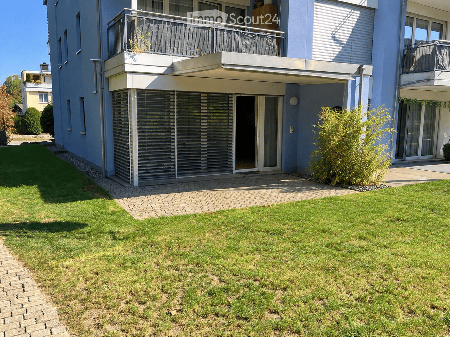 Well-maintained garden with a paved pathway leading to a modern building entrance, featuring balconies and large windows.