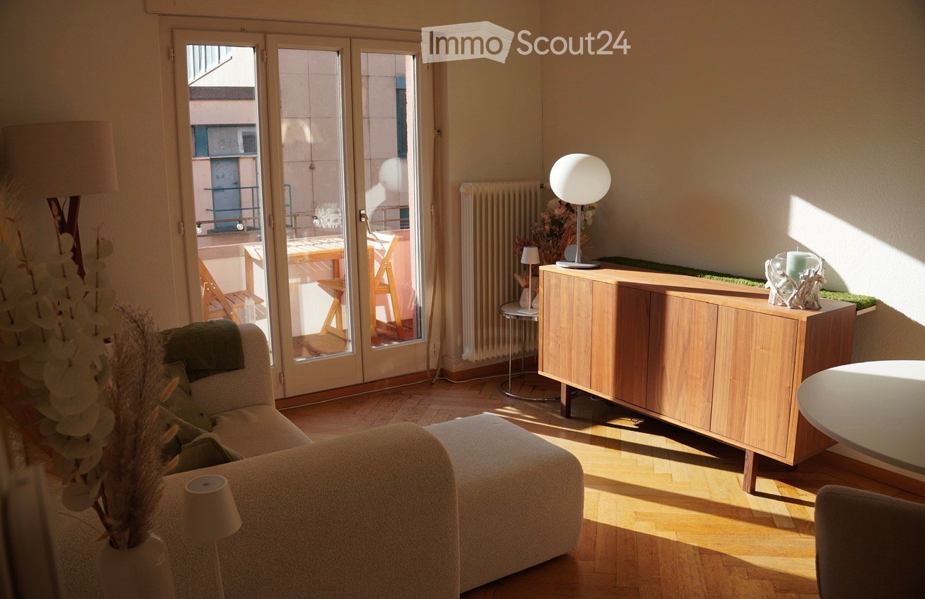 Bright living room with wooden flooring, large sliding glass doors leading to a balcony, a sofa with an ottoman, a coffee table, a sideboard, and decorative lamps.