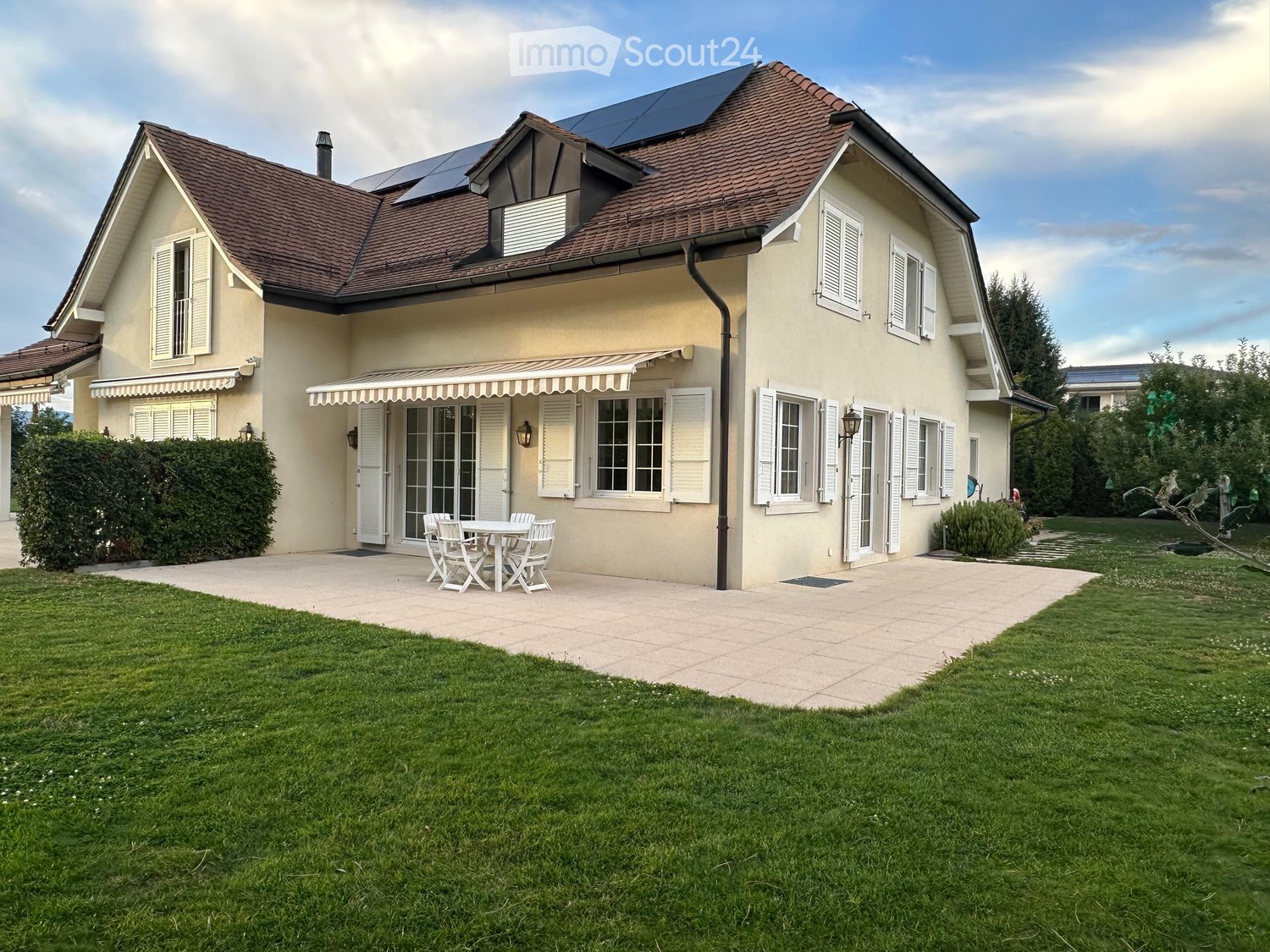 Two-story house with beige walls, brown tiled roof, multiple windows with shutters, solar panels on the roof, and a patio area with a table and chairs.
