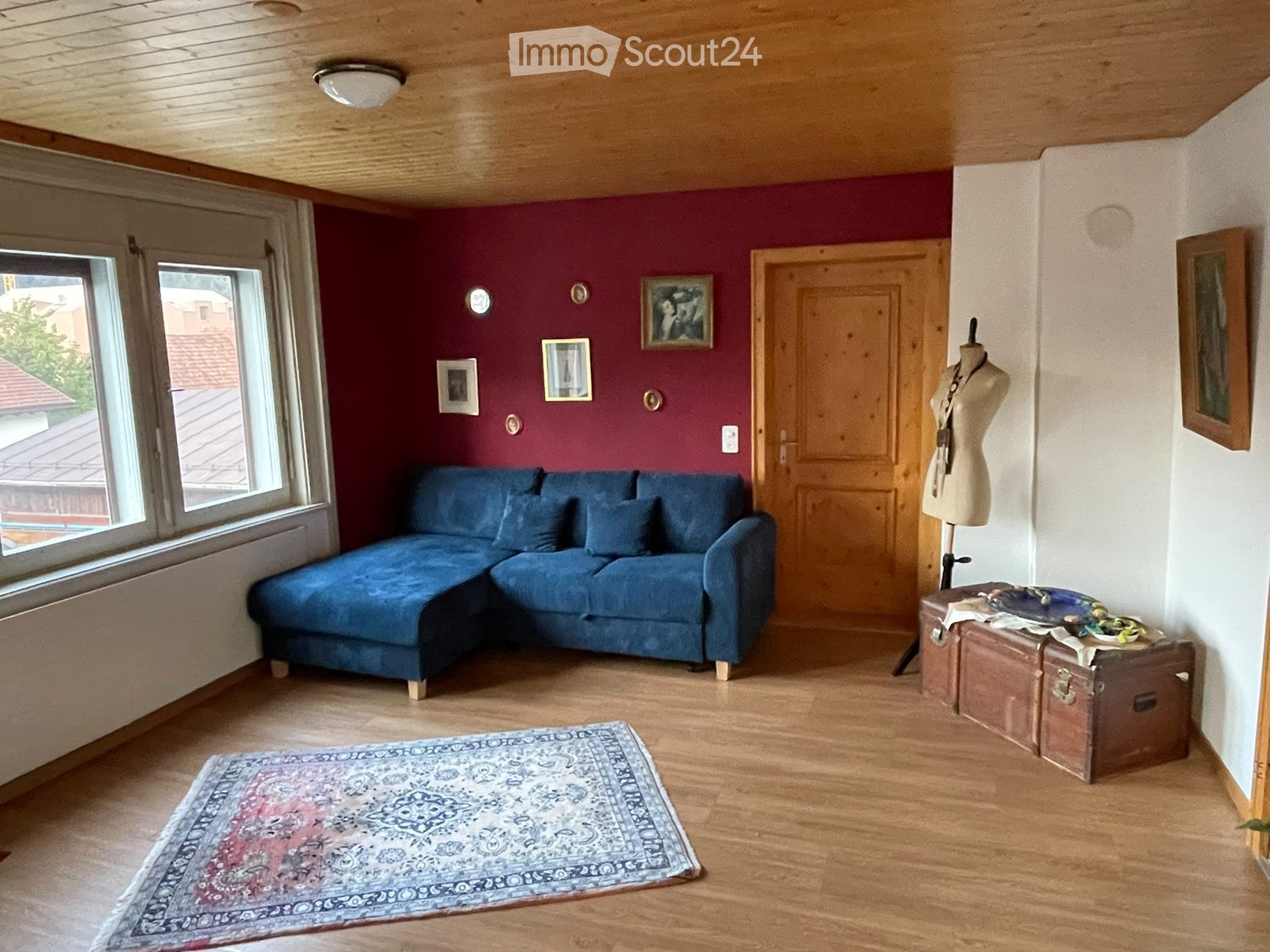 Living room with wooden floors, a blue sectional sofa, a patterned rug, several picture frames on the wall, and a mannequin with accessories on a suitcase.