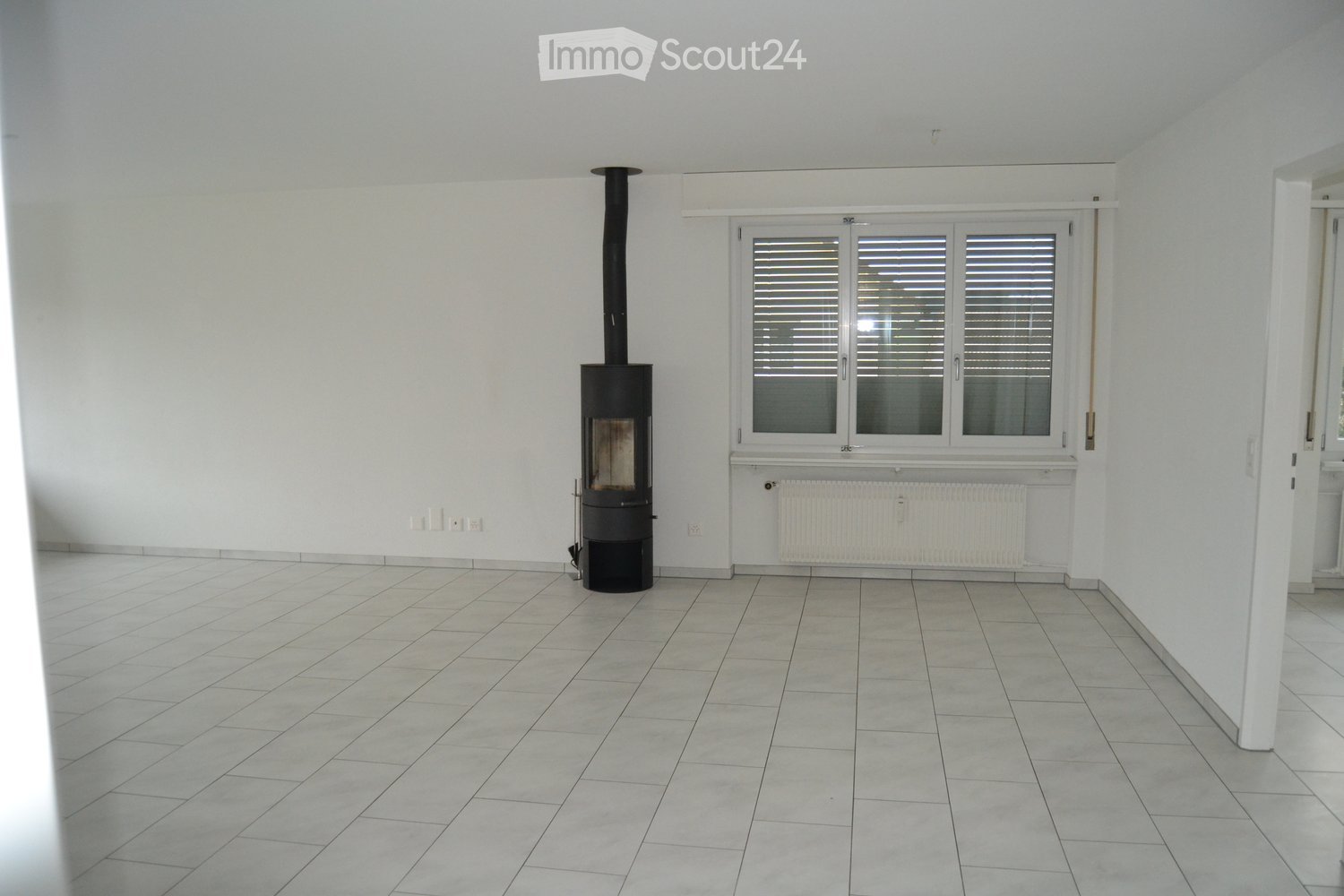 Empty room with tiled floor, white walls, large window with blinds, and a black wood-burning stove with chimney.