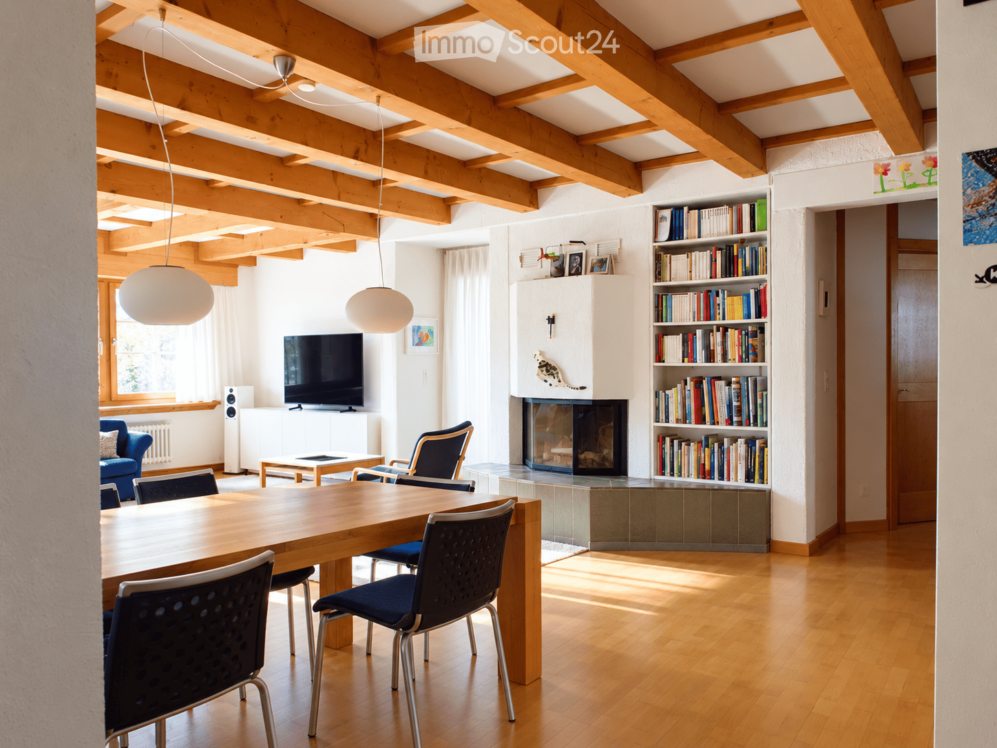 Bright living and dining area with exposed wooden beams, a large wooden dining table with chairs, a blue sofa, a fireplace, a bookshelf, and a television on a white stand.
