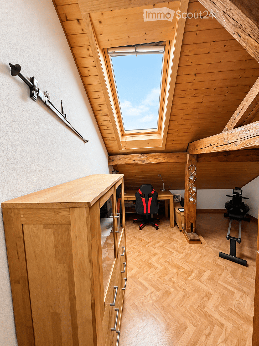 A well-lit attic room with wooden walls and herringbone flooring, featuring a desk with a chair, a cabinet, and an exercise bike.