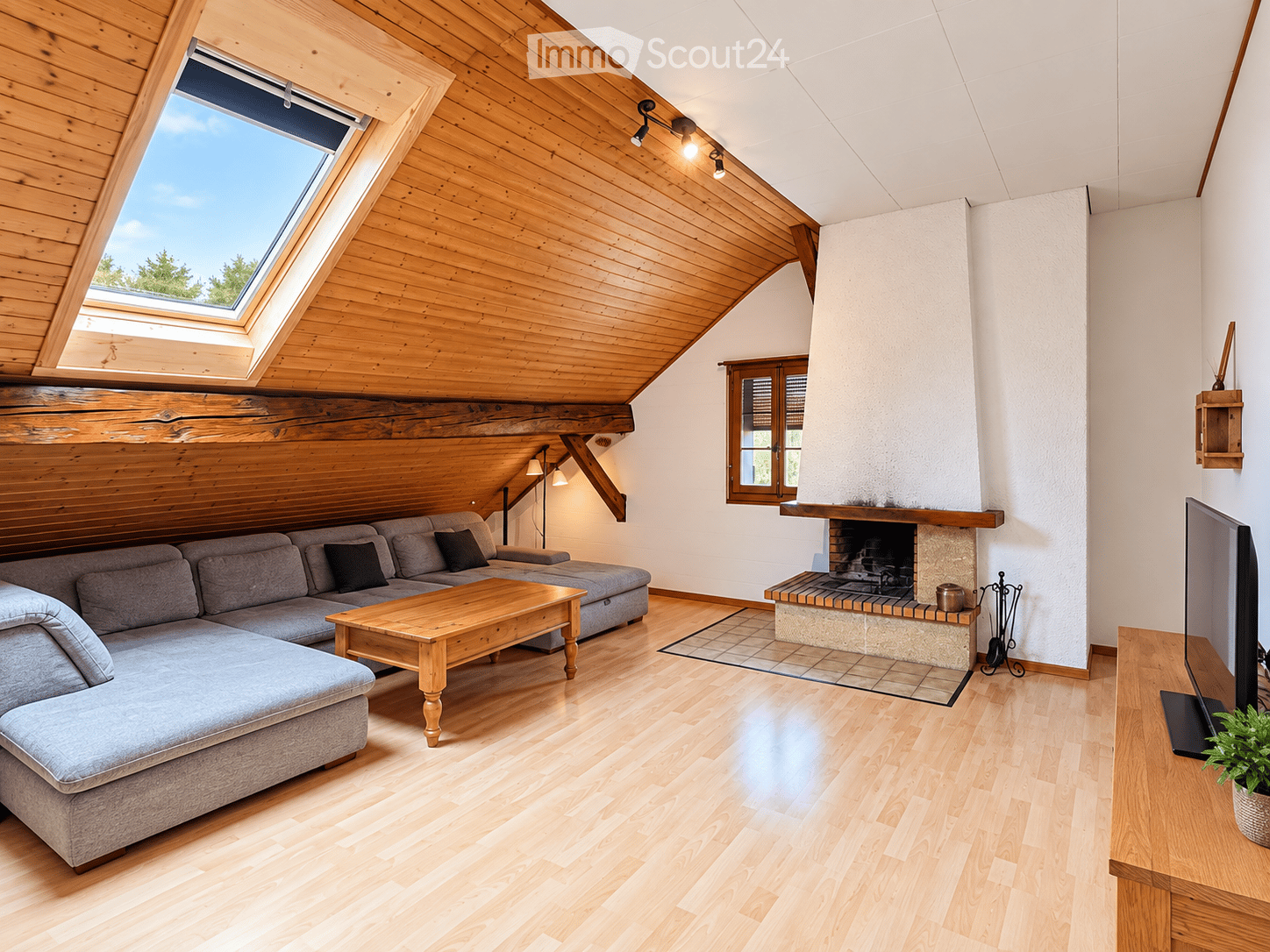 Bright living room with wooden floors, slanted wooden ceiling, skylight window, grey sectional sofa, wooden coffee table, fireplace, TV, and potted plant.
