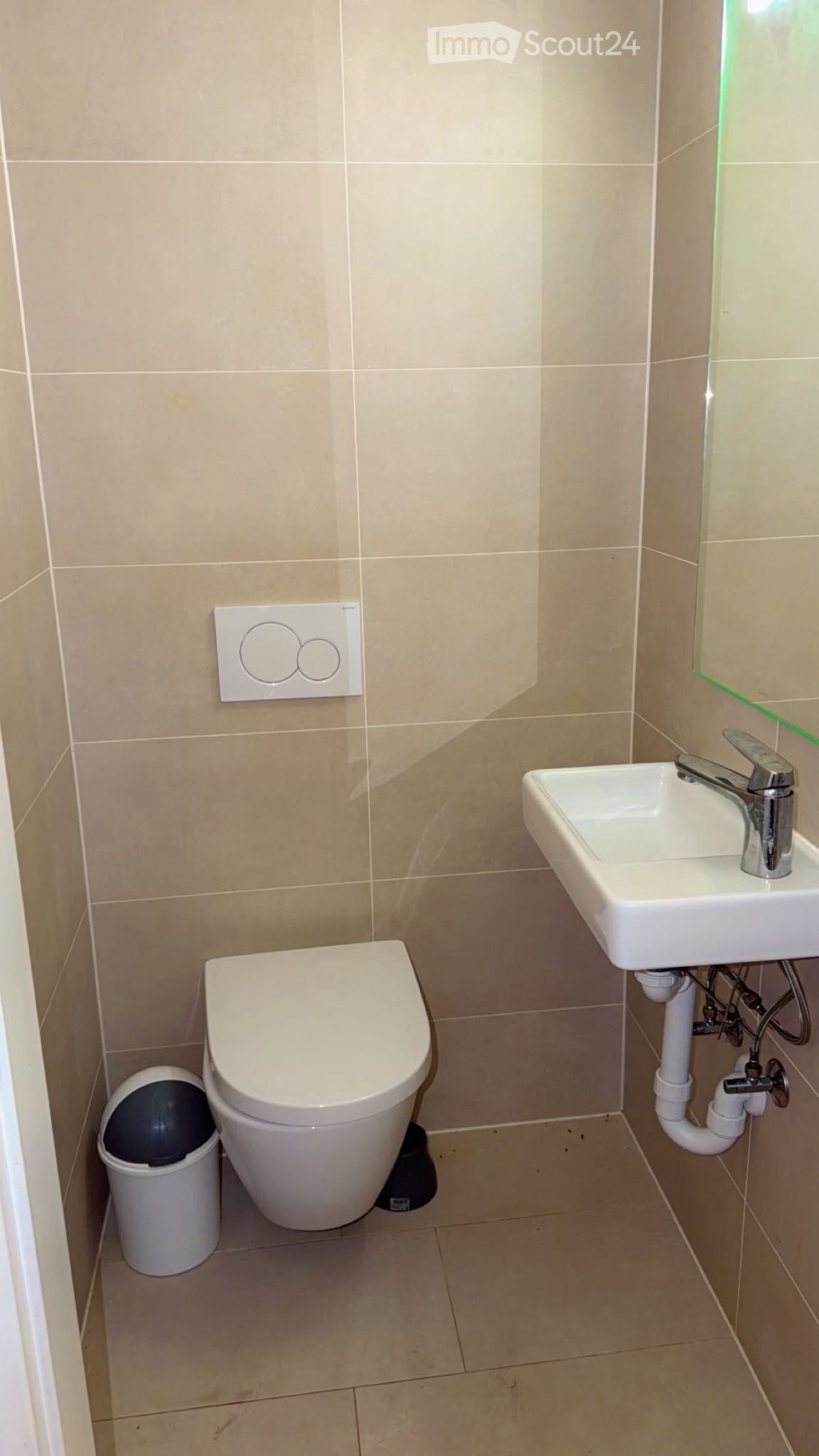 Modern bathroom with beige tiled walls and floor, featuring a toilet, sink with chrome faucet, and a small white trash bin.