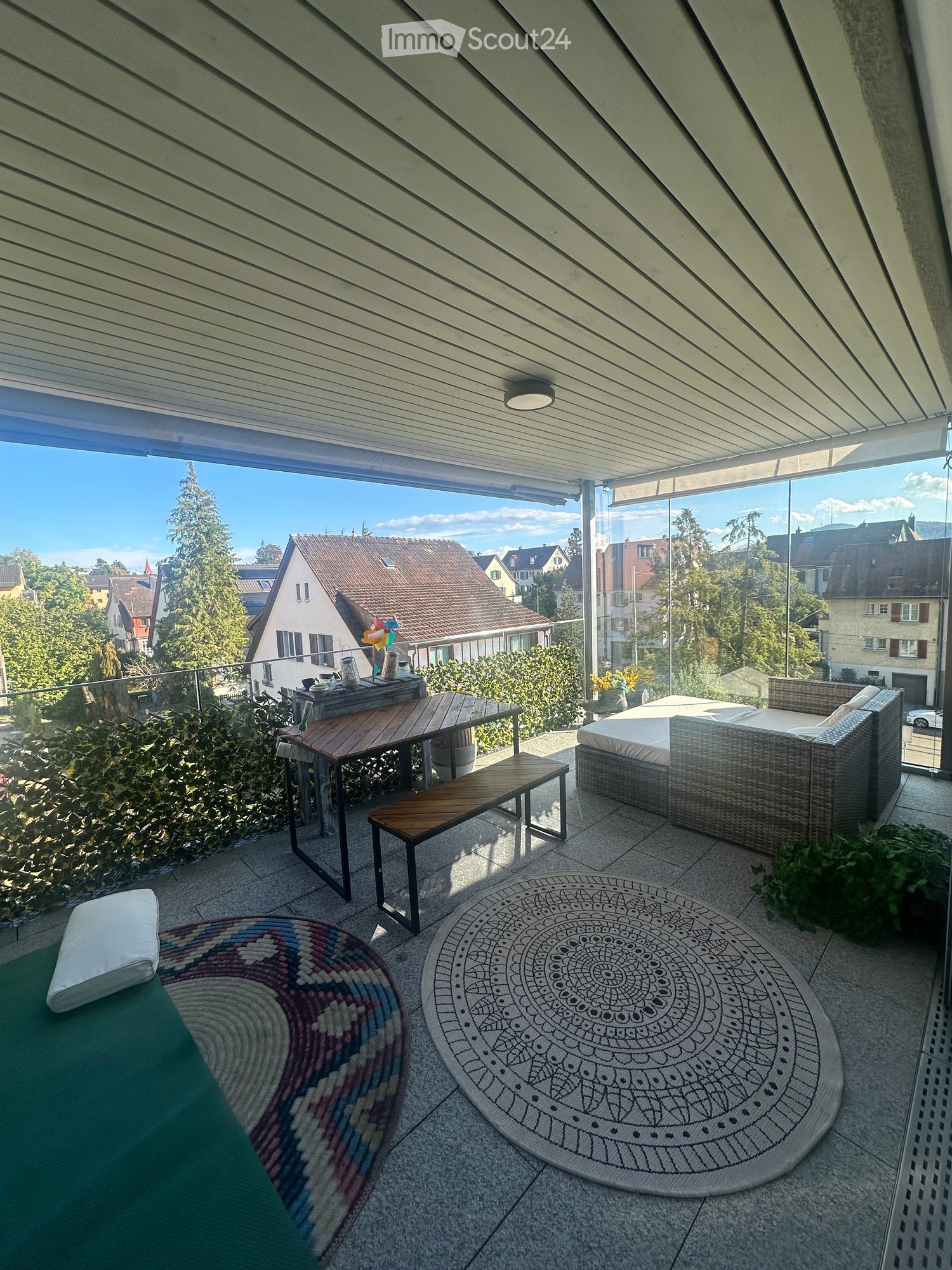 Balcony with a view of the neighborhood, featuring a sofa, a small table, a stool, a bench, and two rugs.