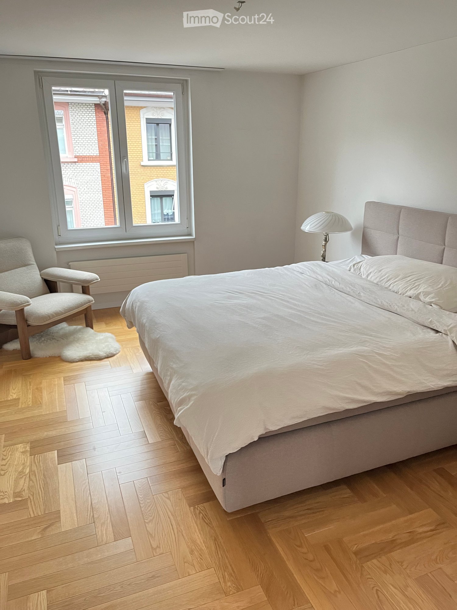 Bright bedroom with wooden flooring, large window, bed with white bedding, armchair, and a lamp on the nightstand.