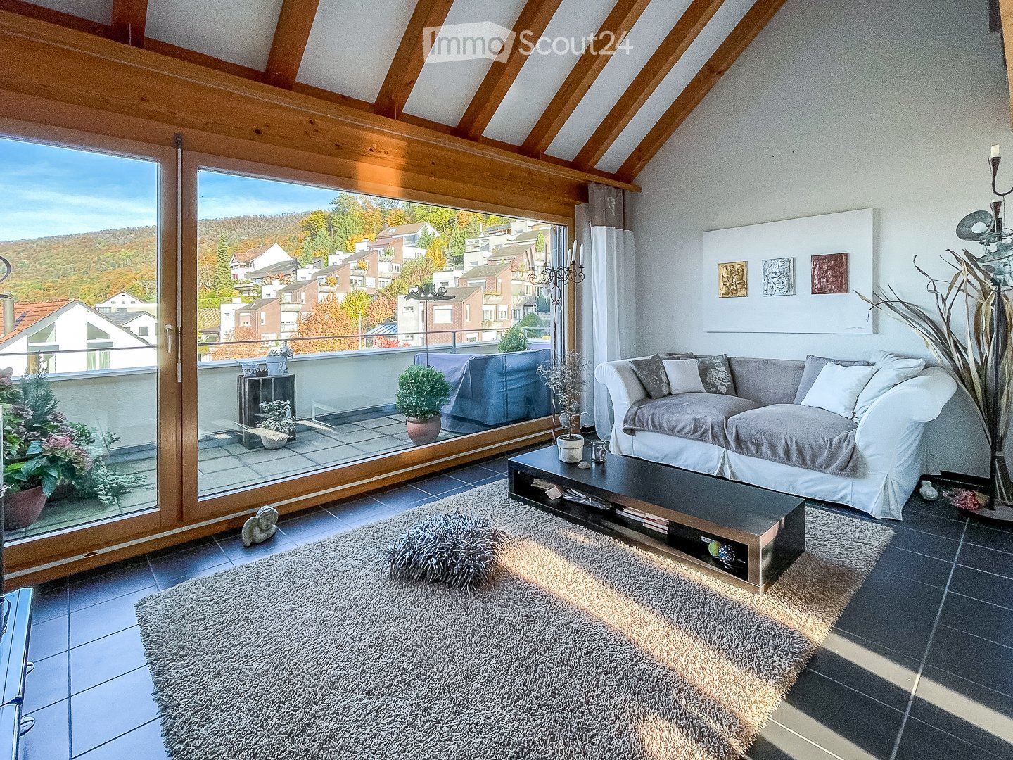 Modern living room with large windows offering a scenic view, featuring a grey sofa, coffee table, shaggy rug, and decorative plants.
