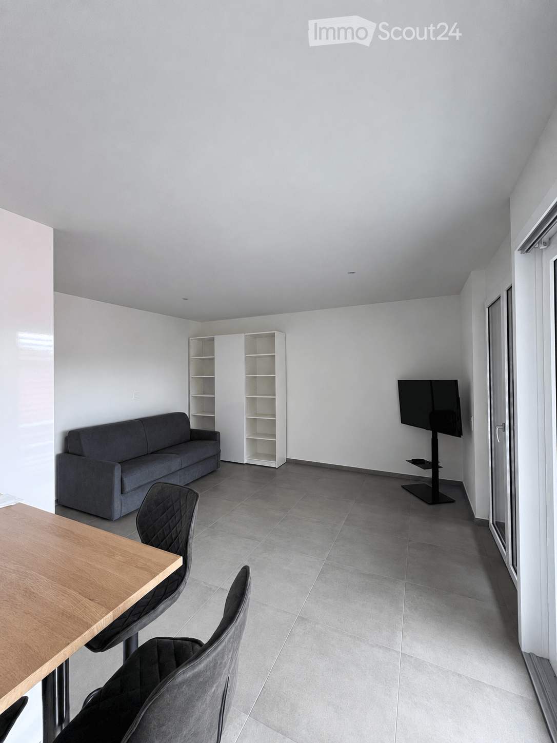 Modern living room with grey sofa, wooden table, chairs, shelves, and a TV on a stand.