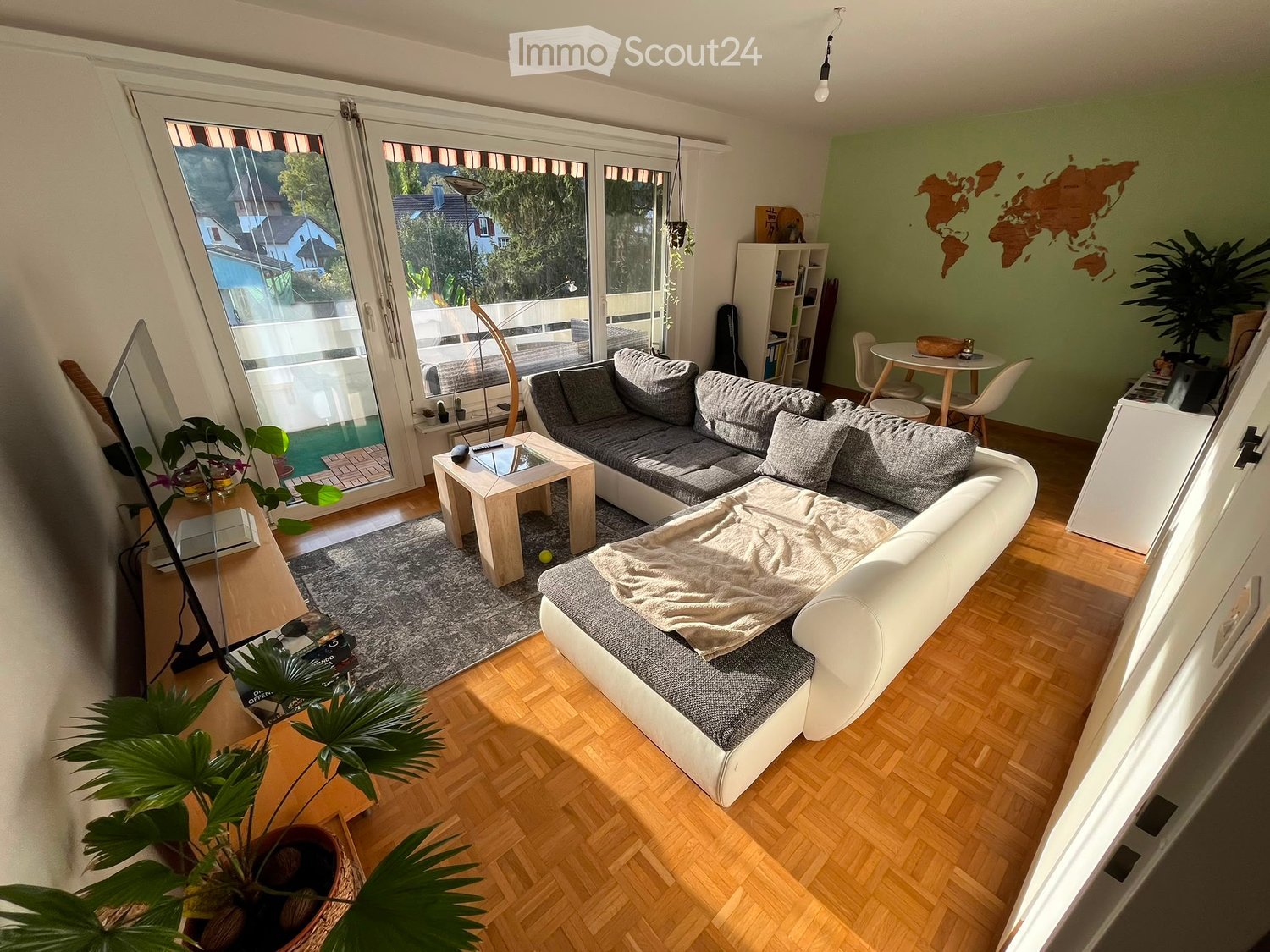 Modern living room with large windows offering a view, featuring a grey sectional sofa, coffee table, dining area with chairs and table, bookshelf, and decorative plants.