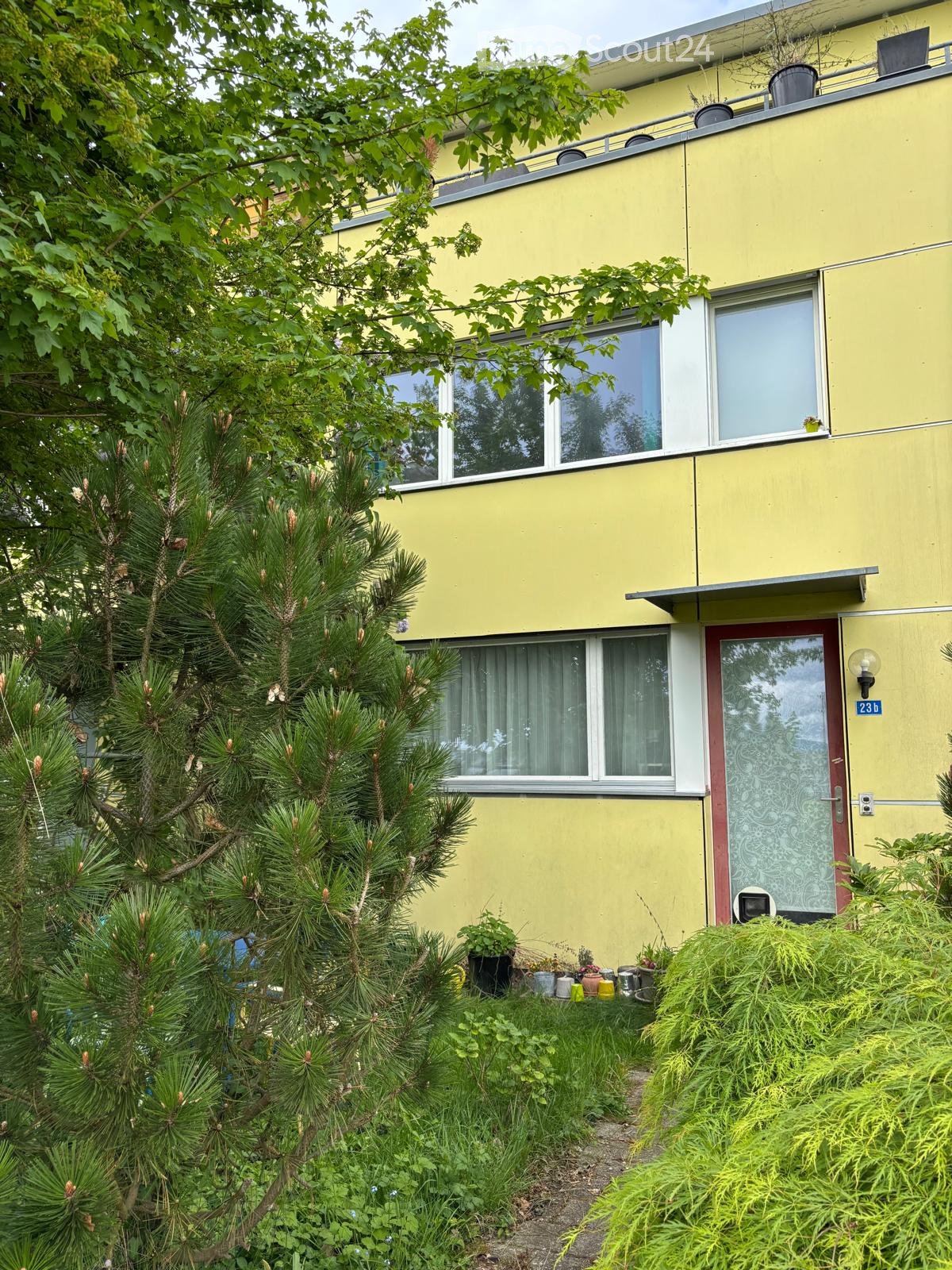 Two-story yellow building with white trim, multiple windows, and a small balcony on the upper floor. The entrance has a red door with a decorative glass panel. The building is surrounded by greenery, including a pine tree and various plants.
