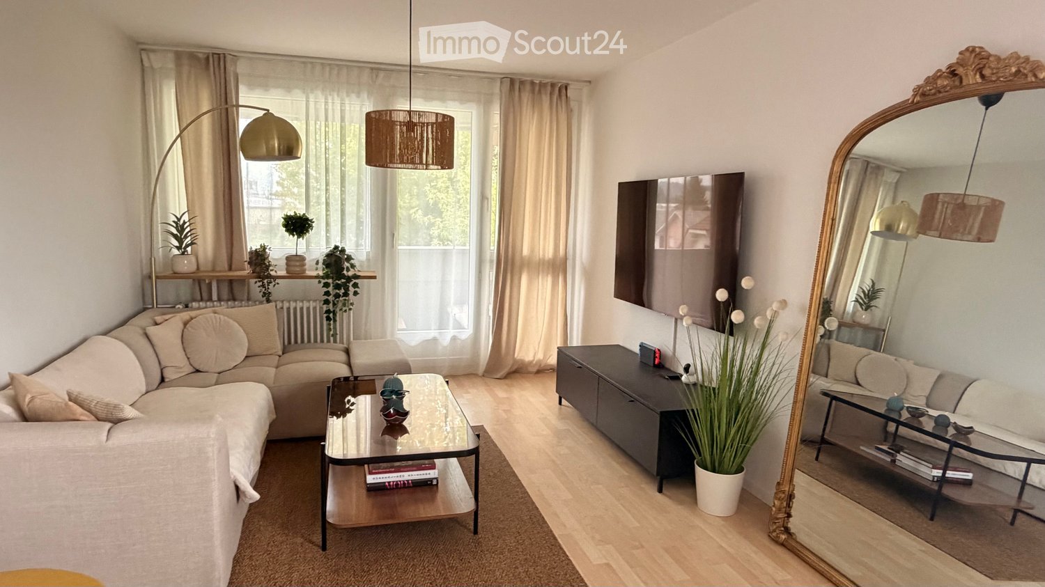 Modern living room with a beige sofa, glass coffee table, large mirror, floor lamp, and decorative plants.