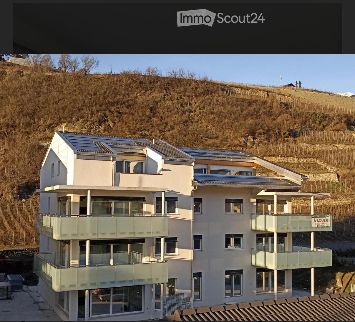 Modern multi-story building with large windows and balconies, situated on a hillside with a clear view of the surrounding landscape.