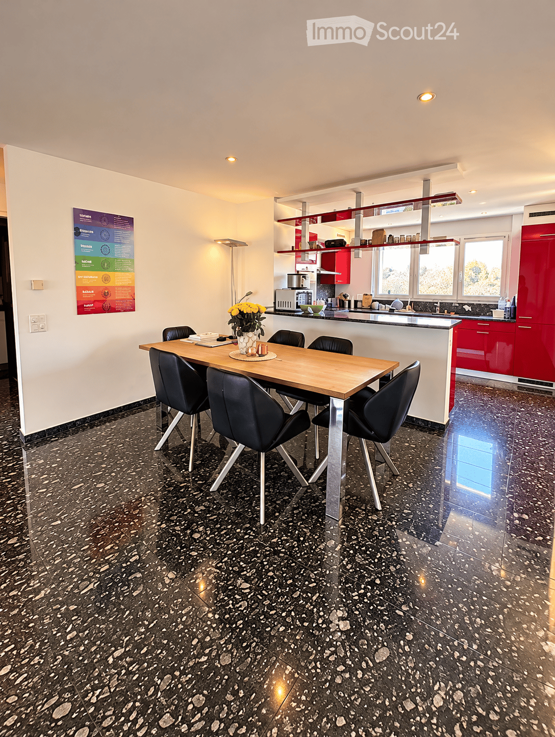 Dining area with a wooden dining table and black chairs, a vase with yellow flowers, and a modern kitchen area with red cabinets, a microwave, and a sink.
